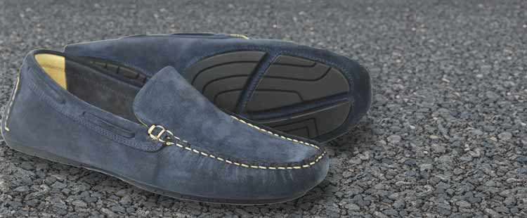 A pair of blue suede loafers with white stitching is placed on an asphalt surface. One shoe is upright, showing the top design, while the other is on its side, revealing the treaded sole.