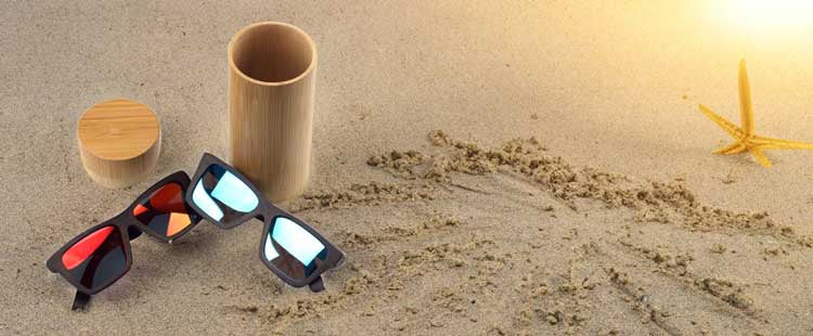 Sunglasses with different colored lenses lie on sand near a bamboo container and a starfish, with sunlight casting a warm glow from the upper right corner.