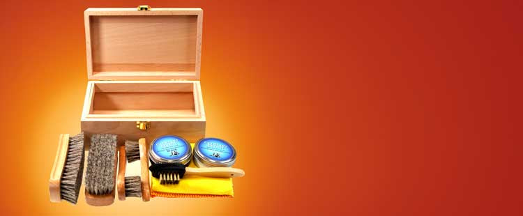 A wooden shoe care kit box is open, surrounded by brushes, tins of polish, a yellow cloth, and an orange cloth, placed on a gradient orange background.