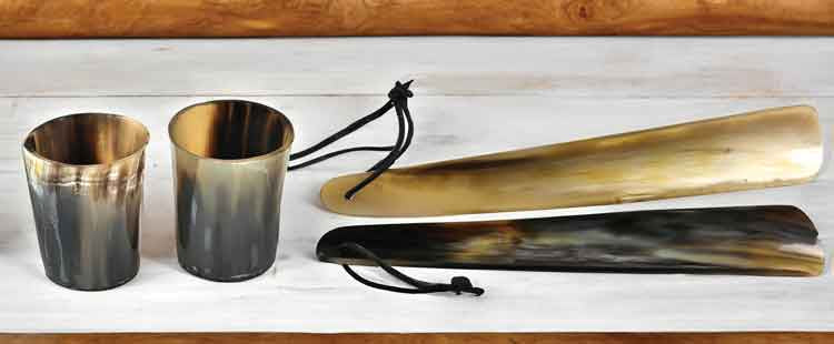 Two horn cups and two horn shoehorns with black leather loops are placed on a light wooden surface, with a textured wooden background. The items display natural variations in color.