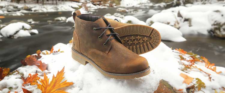 A pair of brown leather boots with black laces on a snow-covered rock, surrounded by autumn leaves, near a flowing stream.