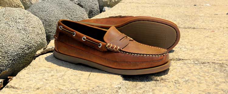 Go to the A pair of brown leather loafers with stitching details and leather laces, placed on stone tiles near large rocks, in bright sunlight. collection