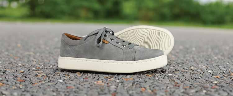 Go to the Gray suede sneakers with white soles are placed on a gravel surface. One shoe stands upright while the other is on its side, showcasing the textured sole. A green, blurred background is visible. collection