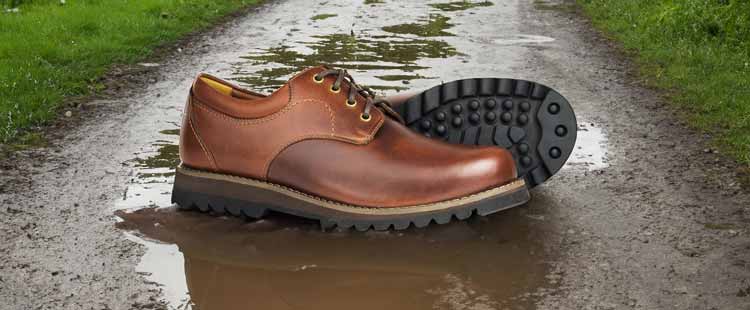 Brown leather shoes with black soles are placed on a muddy path, with green grass on either side. One shoe stands upright, while the other is tilted, showing the textured sole.