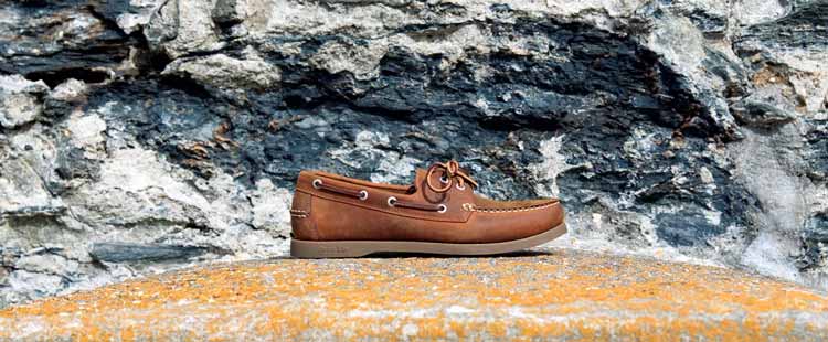 A brown boat shoe with laces sits on a textured stone surface against a rugged stone wall background.