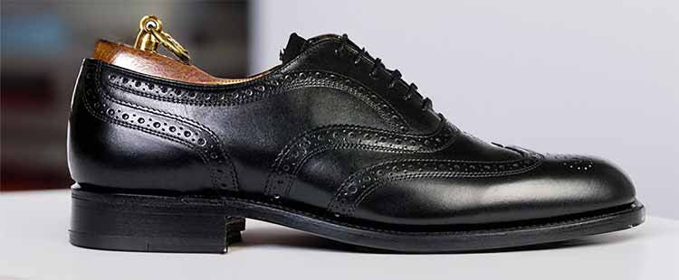 Go to the A polished black leather brogue shoe with detailed perforations and a wooden shoe stretcher inside, placed on a white surface. collection