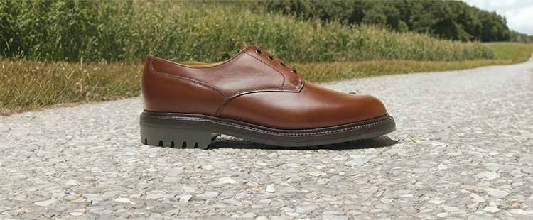 Go to the Brown leather dress shoe with black sole on a gravel path, surrounded by green grass and trees in the background under a partly cloudy sky. collection