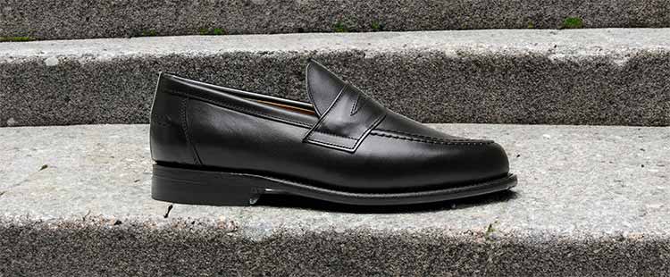 Go to the A black leather loafer is placed on gray stone steps. The shoe features a round toe and a low heel, with visible stitching along the sides. collection