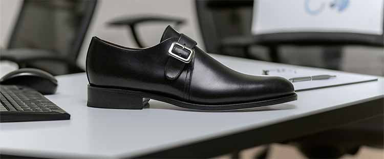 A black leather monk strap shoe with a silver buckle is placed on an office desk next to a keyboard, mouse, and notepad with pens.