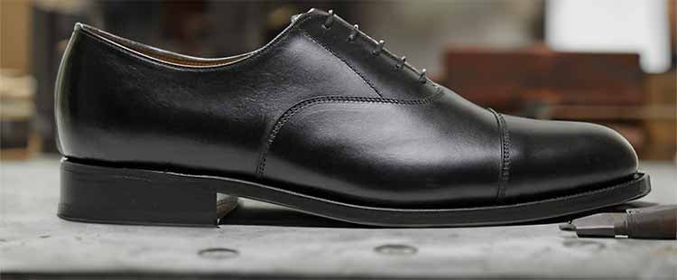 Go to the A black leather oxford shoe with a cap toe design is placed on a gray surface. The shoe features fine stitching and a sleek, polished appearance, showcasing classic formal footwear style. collection