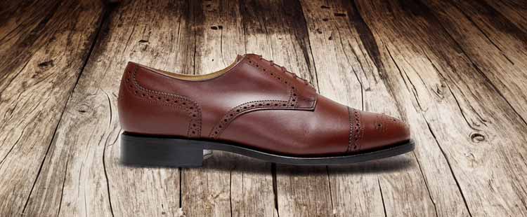 A brown leather dress shoe with intricate brogue detailing is displayed on a rustic wooden floor.