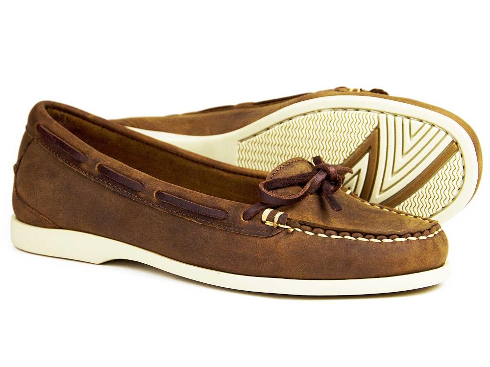 The Orca Bay - Ladies Orca Bay Nubuck Deck Shoe in sand is a moccasin-style loafer with white, non-slip rubber soles. One shoe displays the side profile and decorative lacing, while the other shows off the textured sole. This lightweight design combines style and comfort for any occasion.