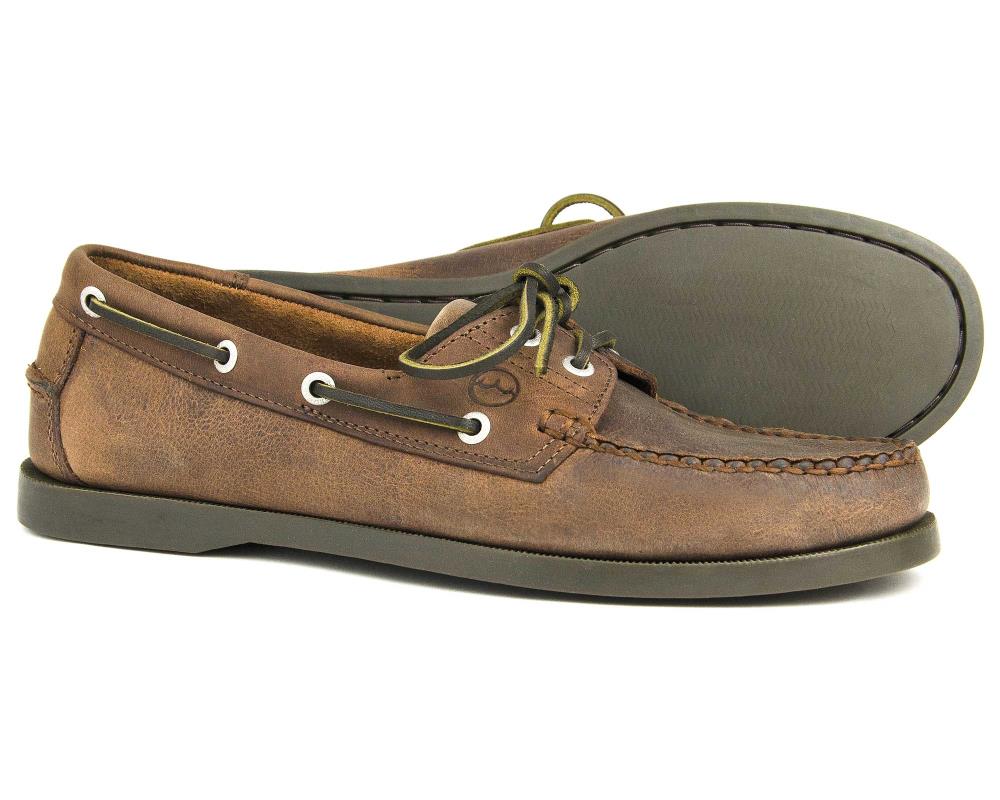 The CREEK Mens Orca Bay Deck Shoe in Russet Brown Nubuck Leather boasts white stitching and lace detailing. Featuring a dark, non-slip rubber sole, one shoe stands upright while the other lies on its side to highlight the durable design.