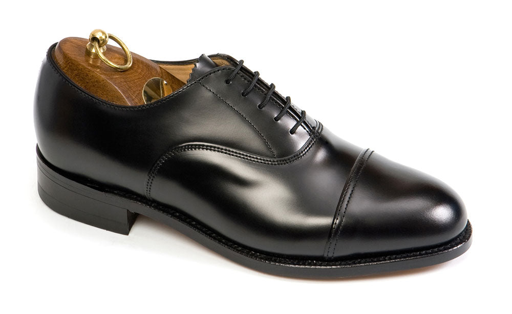 The Sanders OXFORD Extra Wide H Fitting Officers Black Oxford Shoes display sophistication with a polished black leather, a cap toe design, and black laces. Inside, theres a brown wooden shoe tree with a brass handle. The timeless elegance is completed by its low heel.