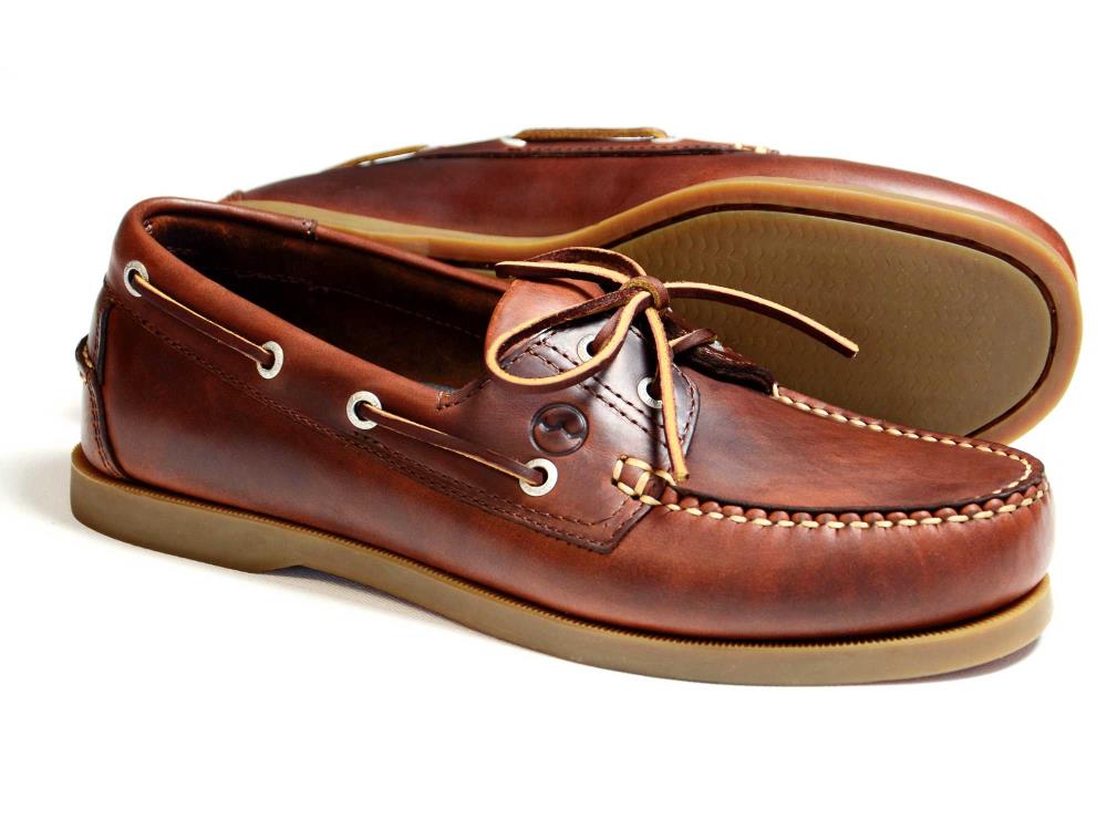 The CREEK Mens Orca Bay Deck Shoe in Saddle Brown features brown leather with white stitching, laced with brown leather string. These shoes have slip-resistant light brown rubber soles and metallic eyelets. One shoe stands upright while the other leans. Brand: Orca Bay.