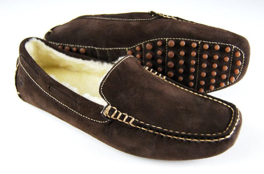 Orca Bays MOHAWK II slippers for men boast a rich brown suede moccasin design, enhanced by visible stitching. They are lined with real sheep fleece for added comfort and feature round rubber grips on the soles, exuding a laid-back elegance with one slipper flat and the other on its side.