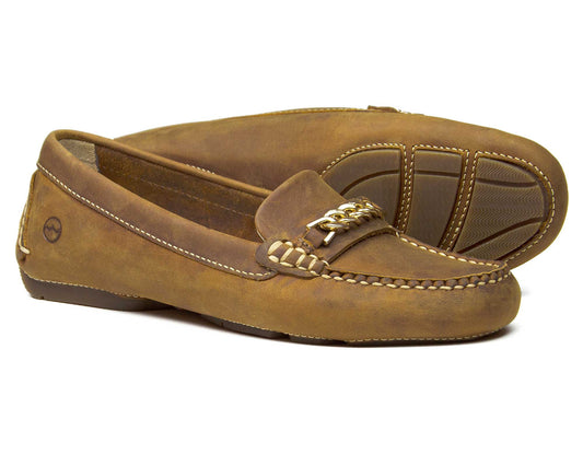 The Orca Bay SALE BADMINTON Womens Sand Nubuck Buckle Loafers feature detailed stitching, a moc toe design, and non-slip rubber soles. They have a decorative metal bit on top, with one shoe upright and the other on its side showing an embossed logo on the heel.