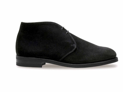 The Sanders CHESHAM black suede chukka boot features a minimalist design with a durable rubber sole, short heel, rounded toe, and sleek Goodyear welted construction. Photographed from the side against a white background, it showcases an elegant profile with classic black laces.