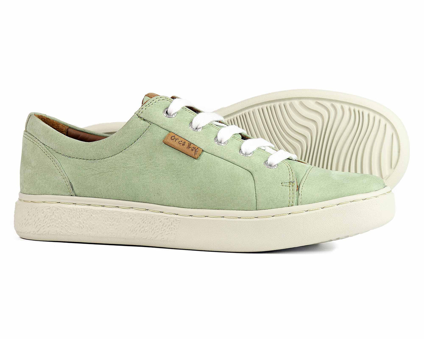 The Orca Bay MAYFAIR Ladies Sneakers in sage green feature white laces and a textured rubber sole. One shoe is upright, the other on its side, showcasing the sole’s pattern. Perfect for summer comfort, a small brown tag is visible on the shoes side.