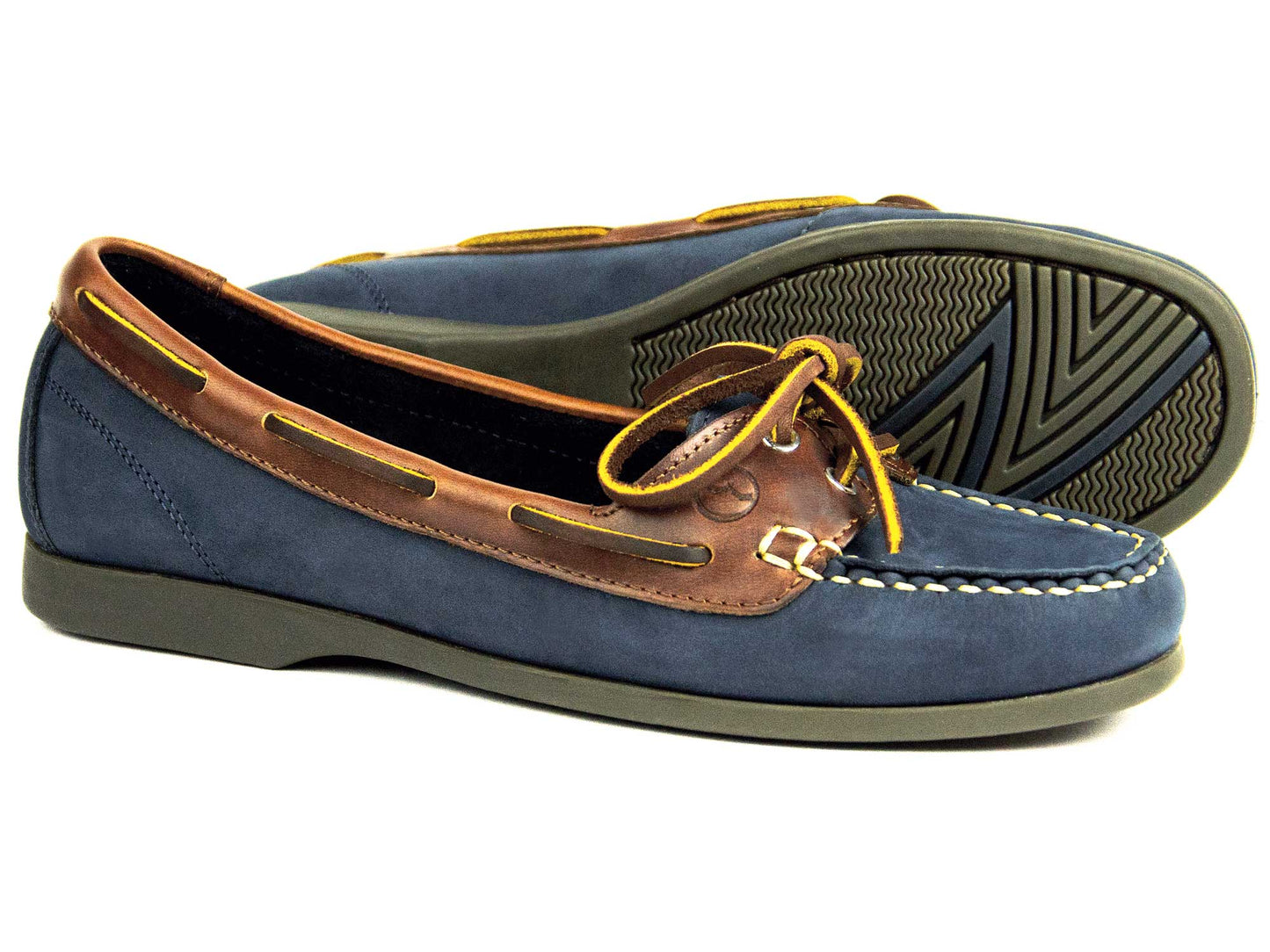 The SCHOONER Ladies Navy Nubuck Deck Shoe by Orca Bay showcases two-tone navy nubuck and brown leather with decorative yellow laces, stitch detailing, and one shoe displayed upright while the other is on its side to highlight the treaded sole.